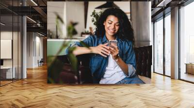 Relaxed young african woman texting on her phone in the modern house.Smiling african american woman using smartphone at home, messaging or browsing social networks while relaxing on couch Wall mural