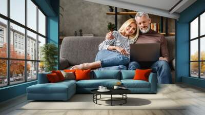 Senior couple using laptop and smiling while resting on couch at homewhile watching movie Wall mural