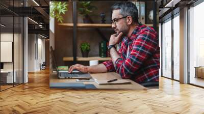 Side view shot of pensive young man sitting at home office and working on laptop Wall mural