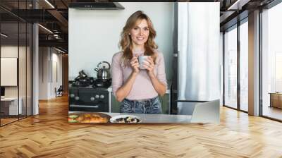 Smiling Caucasian woman lady with blond hair casual outfit standing in the modern kitchen having eating healthy breakfast holding cup mug drink hot coffee tea using laptop looking at camera Wall mural