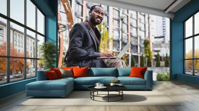 Smiling confident 30s african man using laptop with cup of coffee in city area. Young male freelancer ceo manager working remotely, typing, doing projects startup outdoors Wall mural