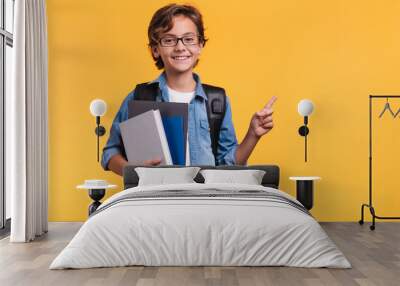 Smiling little boy pointing at copy space in casual clothes with books for studing at school isolated over yellow background Wall mural