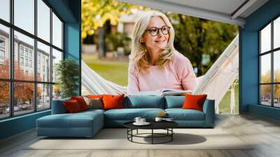 Smiling senior good-looking blond woman wearing glasses while reading in hammock in the summer garden Wall mural