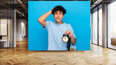 Surprised boy guy high school male pupil student teenager with dental braces backpack holding alarm clock isolated over blue background. Time management education back to school concept Wall mural