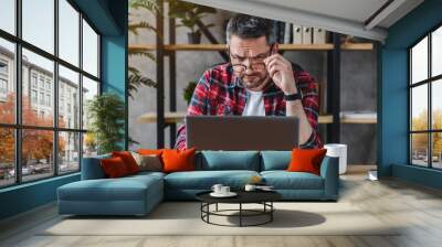 Thoughtful businessman in eyewear looking at computer screen, sitting at table at home Wall mural