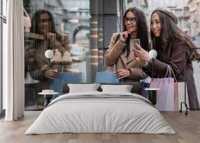 Two happy girlfriends looking on the shopwindow while photographing on smartphone with shopping bags near the mall Wall mural