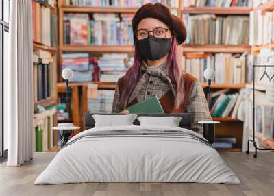 Young caucasian clever hipster high school girl pupil college student teenager wearing medical mask to protect from virus holding a book in the university library or bookshop. Education concept Wall mural