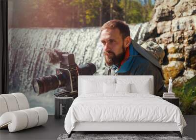 Travel photographer bearded man close-up with professional film camera on tripod shooting mountain landscape in waterfall background. Hiker tourist professional photography shooting, filming backstage Wall mural