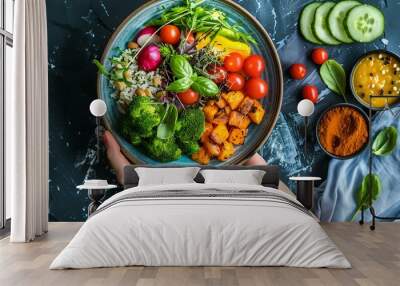 A person is holding a blue bowl filled with a variety of vegetables, including broccoli, carrots, and tomatoes. The bowl is placed on a counter next to a plate of vegetables and a bowl of sauce Wall mural