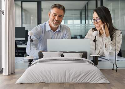 2 multicultural team coworkers, older Latin man executive and young Asian woman manager working together, happy diverse employees looking at laptop computer discussing financial corporate project. Wall mural