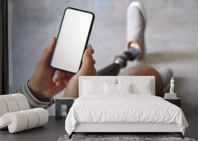 Amputee man with disability leg prosthesis sitting on sofa at home holding cell using mobile phone with big blank white empty screen mock up template for advertising concept. Close up Wall mural