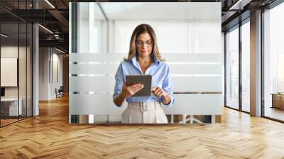 Busy middle aged businesswoman manager wearing eyeglasses looking at tablet standing in office. Mature professional business woman executive using tab, female entrepreneur or hr working with pa. Wall mural