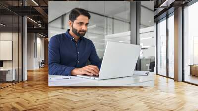 Busy professional Indian business man employee looking at laptop computer, businessman office worker or executive manager analyzing project strategy working online with ai software at work desk. Wall mural