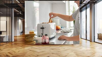 Close up view of teenage boy cooking in the kitchen, holding bread slice in hand making toasts in toaster for snack preparing family breakfast or lunch alone at home. Wall mural