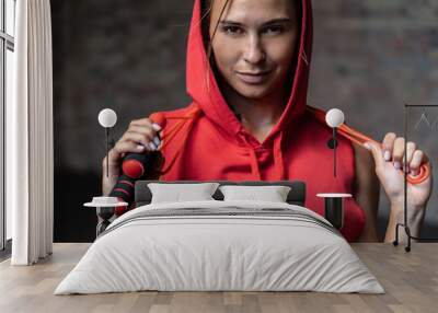 Closeup portrait pretty smiling sporty young woman in red hoody sportswear hold skipping jumping rope around neck happy look at camera brick wall background weightloss cardio healthy,lifestyle concept Wall mural