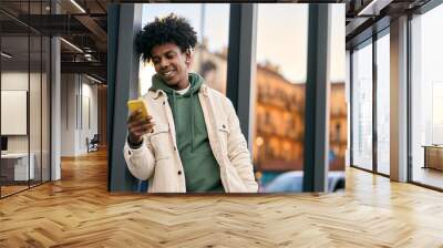 Cool smiling young African American teen guy holding mobile phone standing at glass city wall. Happy stylish hipster teenager tech user using cell outdoors, looking at cellular checking smartphone. Wall mural