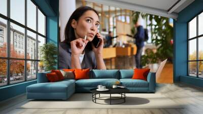 Doubtful Asian young adult business woman talking on cell phone making call working looking away thinking of solution sitting in cafe, considering offer, making decision or feeling doubt. Wall mural