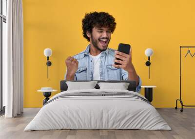 Excited happy young indian man winner feeling joy using smartphone winning lottery game, betting, getting cashback online gift in mobile app message holding cell phone isolated on yellow background. Wall mural