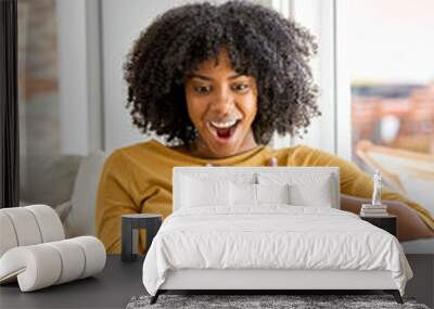 Excited young African American woman customer looking at mobile phone sitting on sofa at home. Happy lady using smartphone surprised about money win, lottery prize, shopping cashback or gift. Vertical Wall mural