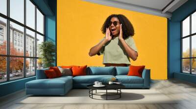 Excited young Black woman wearing sunglasses standing at yellow wall. Happy surprised African American gen z girl wow face feeling surprise looking away advertising summer shopping sale, travel offer. Wall mural