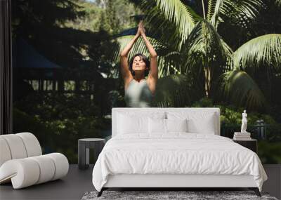 Fit sporty mindful young Hispanic woman meditating doing yoga breathing exercises with eyes closed feeling peace of mind, mental balance standing in green nature tropical park on sunny day. Wall mural