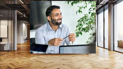 Friendly happy smiling ceo Indian businessman holding pen in hand looking listening to colleague discussing global corporation research report at table. Executive manager working in modern office. Wall mural