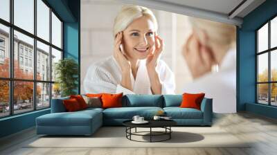 Gorgeous mid age adult 50 years old blonde woman standing in bathroom after shower touching face, looking at reflection in mirror smiling doing morning beauty routine. Older dry skin care concept. Wall mural