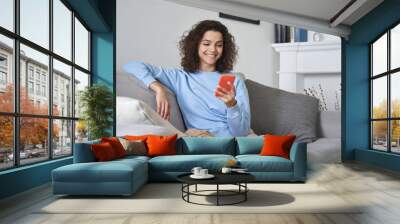 Happy 20s young woman watching social media videos looking at smartphone relaxing on couch, smiling hispanic girl enjoying using online mobile apps doing online shopping on cell phone at home. Wall mural