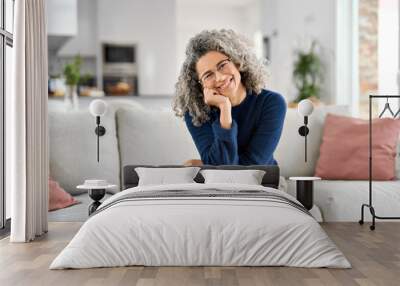 Happy 50 years old woman sitting on sofa at home looking at camera. Smiling middle aged lady posing in living room at home. Mature older female with curly gray hair relaxing on couch. Portrait. Wall mural