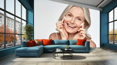 Happy 50s mid aged mature old blonde lady applying facial cream on face skin laughing enjoying anti age fresh soft skin care beauty spa treatment skincare creme isolated on white background. Portrait Wall mural