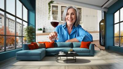 Happy 60s mature woman drinking wine video calling friend at home. Old middle aged lady holding glass talking to web cam by online social distance chat meeting sit at kitchen table. Webcam view. Wall mural