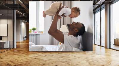 Happy affectionate young Black dad lifting adorable mixed race baby daughter having fun in bed at home. Smiling African father playing with cute funny infant child girl waking up in bedroom in morning Wall mural