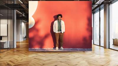 Happy African American teen guy having fun laughing on red wall lit with sunlight. Smiling cool ethnic gen z teenager model standing looking at camera, authentic full length shot outdoors. Wall mural