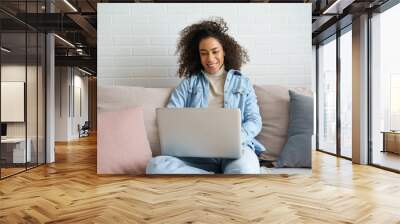 Happy african teen girl student remote worker using laptop computer sit on sofa at home office. Young black woman surfing internet, communicate online in social media, video calling, watching movie. Wall mural