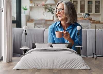 Happy beautiful relaxed mature older adult grey-haired woman drinking coffee relaxing on sofa at home. Smiling stylish middle aged 60s lady enjoying resting sitting on couch in modern living room. Wall mural