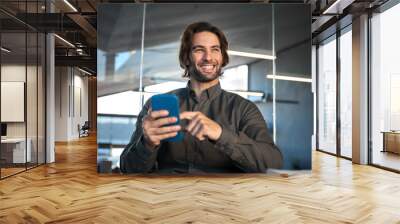 Happy busy smiling young business man executive, businessman manager user sitting in office holding smartphone using mobile cell phone doing bank financial payment on cellphone looking away at work. Wall mural