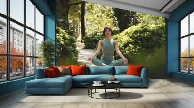 Happy calm mindful young Hispanic woman meditating doing yoga breathing exercises with eyes closed feeling peace of mind, mental balance sitting in lotus pose in green nature tropical park outdoors. Wall mural