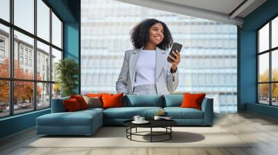 Happy confident young African American business woman using cell mobile phone outdoor. Smiling lady professional holding cellphone standing at city street building looking away working outside office. Wall mural