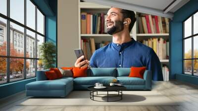 Happy ethnic indian professional business man at work in home office wearing earbud using cell phone, smiling eastern businessman having virtual mobile chat video call on smartphone, watching game. Wall mural