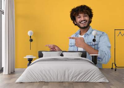 Happy excited young indian man looking at camera pointing aside with fingers hand gesture at copy space advertising new promotion, presenting sale offer standing isolated on yellow background. Wall mural