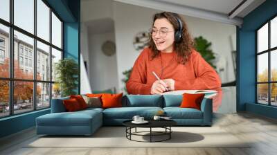 Happy female student wearing headphones elearning online on computer at home. Young smiling woman using laptop remote working, talking having web conference meeting, virtual class, web seminar. Wall mural