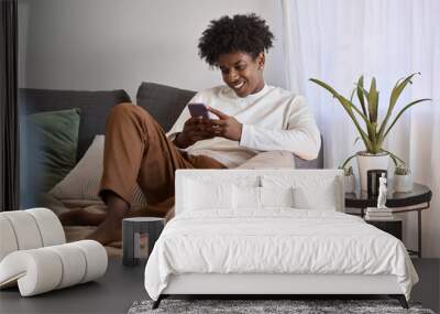 Happy gen z African American teen sitting on couch holding smartphone, using cell phone, looking at cellphone enjoying doing online ecommerce shopping in mobile apps, chatting or playing games. Wall mural