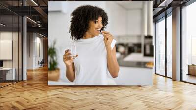 Happy healthy young adult African American woman model talking pill holding glass of water standing in the kitchen at home. Morning supplements vitamins nutrition treatment concept. Wall mural