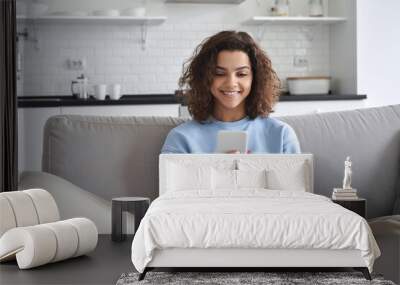 Happy hispanic teen girl looking at smartphone relaxing on couch at home, enjoying using online mobile apps technology, playing games on cell phone, checking messages or social media posts. Wall mural