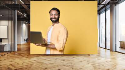 Happy indian business man student standing isolated on yellow background holding laptop advertising online products for learning, education training and webinars, study video tutorial course. Wall mural