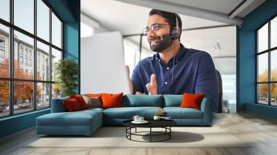 Happy Indian call center agent wearing headset talking to client working in customer support office. Professional contract service telemarketing operator using laptop having conversation. Candid shot Wall mural