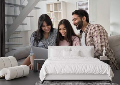 Happy indian family with child daughter having fun using laptop computer at home. Smiling parents and teen kid daughter laughing looking at device browsing or watching funny videos sitting on sofa. Wall mural