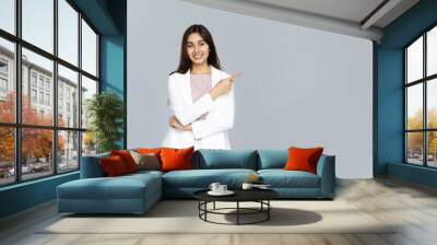 Happy indian young business woman wear white suit looking at camera pointing finger at copy space isolated on grey studio background, smiling beautiful confident hindu lady professional showing aside Wall mural