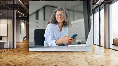 Happy mature business woman executive holding mobile cell phone looking away in office. Smiling middle aged professional businesswoman manager wearing glasses using cellphone working on smartphone. Wall mural