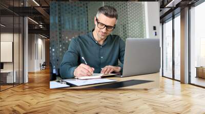 Happy mature executive ceo manager using laptop writing notes in notebook at workplace. Smiling middle aged business man working in office analyzing financial data doing research. Wall mural